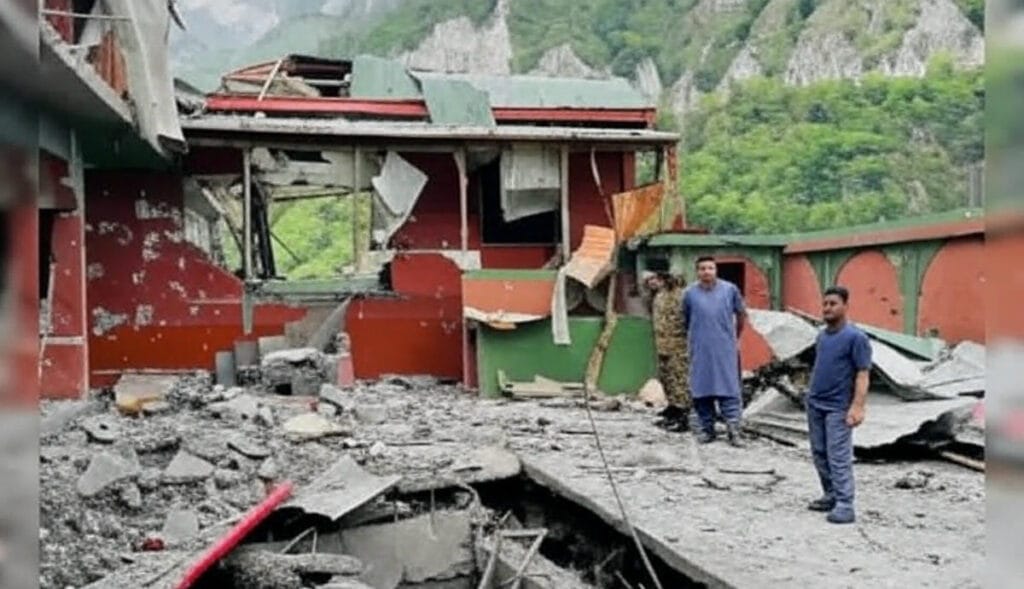 Two people stand amidst the rubble of a heavily damaged building in a mountainous area, with debris including broken concrete, metal sheets, and wooden beams scattered around. The structure, painted red and green, has collapsed sections and exposed interiors, with lush green mountains and rocky cliffs in the background. The scene suggests the aftermath of a destructive event.