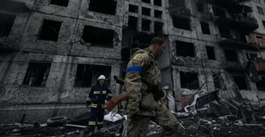 A scene of devastation featuring a heavily damaged multi-story building with broken windows and exposed interiors. In the foreground, a soldier in camouflage uniform with a blue armband walks forward, carrying a rifle. Behind him, a firefighter in a reflective uniform stands near debris, holding an axe. The ground is littered with rubble, bricks, and a tire, indicating recent destruction.