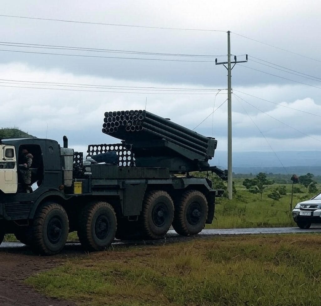 The image depicts a military truck carrying a multiple rocket launcher system traveling on a rural road. The truck, painted in green and beige camouflage, features multiple rocket tubes mounted on its rear. A soldier is visible in the driver's cabin. The scene is set against a backdrop of grassy fields, a cloudy sky, and a utility pole with power lines. A civilian car is seen on the right side of the road, suggesting ongoing activity in the area.