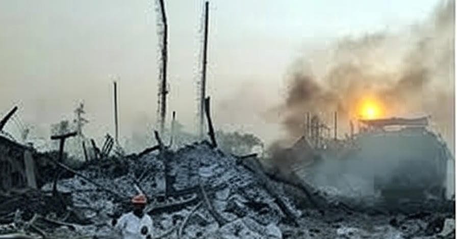 The image depicts a scene of significant destruction, featuring a large pile of rubble and debris in the foreground, with a person wearing a white shirt and an orange cap standing amidst the wreckage. In the background, a structure is engulfed in flames and thick smoke, with the sun partially visible behind it, casting an orange glow. The sky is hazy due to the smoke, and several charred poles or beams protrude from the debris.