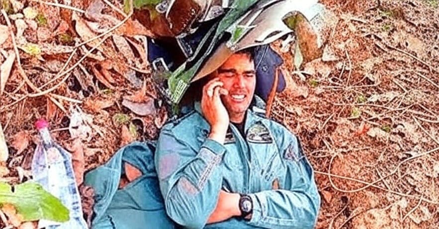 A pilot in a green flight suit lies on the ground amidst wreckage and scattered leaves, holding a phone to his ear, with a water bottle and debris nearby in a forested outdoor setting.