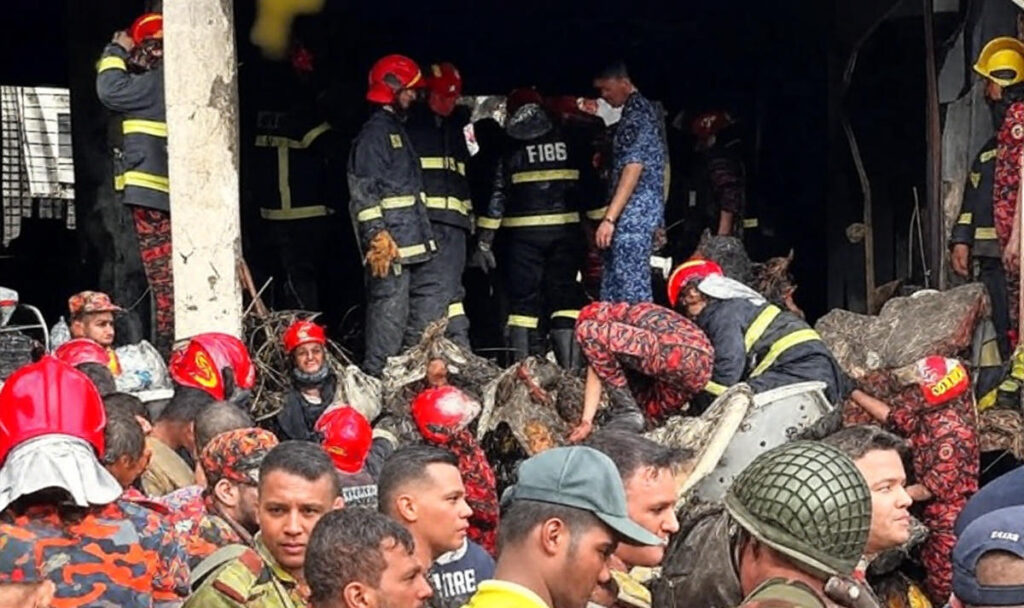 Emergency responders, including firefighters and military personnel, work together amidst the wreckage inside a heavily damaged building, with firefighters in red helmets and dark uniforms navigating debris, following the Bangladesh Air Force jet crash on July 21, 2025, in Dhaka.