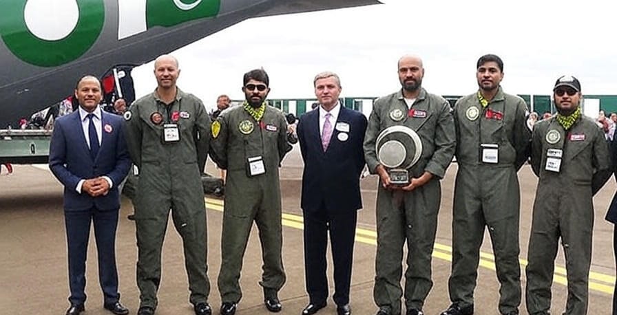 A group of individuals stands in front of a Pakistan Air Force C-130 Hercules aircraft with a green and white "PAF" livery. The group includes five personnel in green flight suits adorned with badges and name tags, two individuals in formal suits with name badges, and one person holding a trophy. The scene takes place on an airfield with a cloudy sky in the background, and yellow taxiway lines are visible on the ground.
