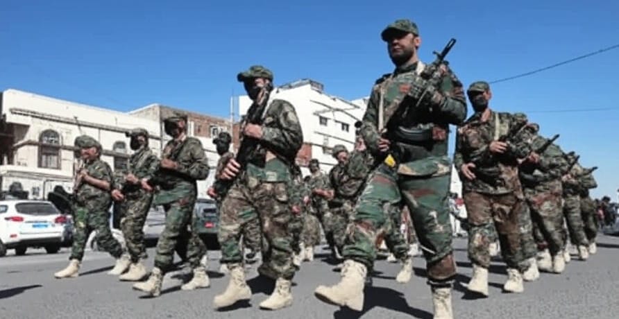 The image depicts a group of soldiers in camouflage uniforms marching in unison on a paved street under a clear blue sky. The soldiers are equipped with rifles and wear tactical gear, including caps and beige combat boots. They are moving in a disciplined formation, with buildings and a white vehicle visible in the background, suggesting an urban setting. The scene appears to be part of a military parade or exercise.