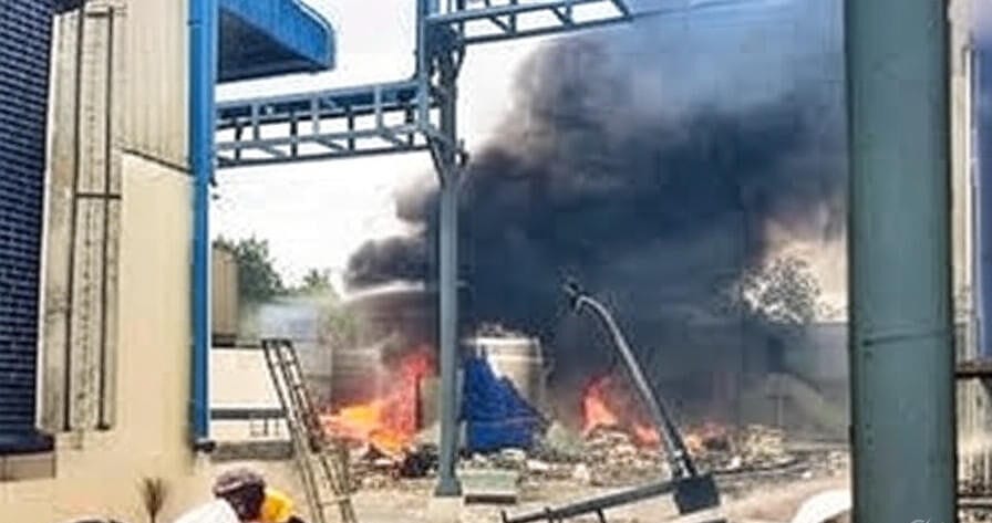 Emergency responders in protective gear combat a massive fire and thick black smoke billowing from a damaged industrial facility, with flames engulfing a chemical storage unit and debris scattered around the site. Fire hoses are actively deployed, while personnel assist in the chaotic scene, framed by the structure's blue and yellow exterior and overhead framework.