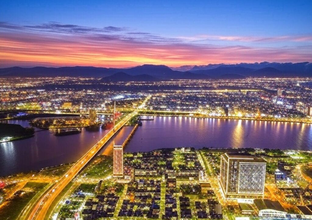 The image shows a breathtaking aerial view of a cityscape at dusk, with a vibrant sunset sky transitioning from deep blue to warm orange and pink hues. A river runs through the center, reflecting the city lights, and a brightly illuminated bridge stretches across it, connecting the two sides. The city is filled with a mix of modern high-rise buildings and residential areas, with lights twinkling against the backdrop of distant mountains. The scene captures a blend of urban development and natural beauty, with visible roads and a sports field in the foreground.
