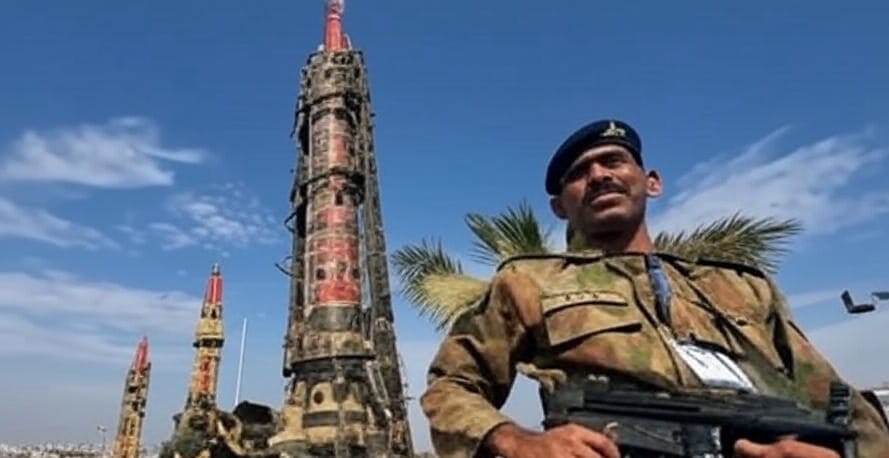 An image depicting a military scene with a soldier in camouflage uniform and a beret holding a submachine gun in the foreground. Behind the soldier, there is a large, upright missile adorned with red and black markings, flanked by additional missile structures. Palm trees are visible in the background, along with a clear blue sky, suggesting a location with a warm climate. The setting appears to be a military display or parade, emphasizing the presence of advanced weaponry and military personnel.