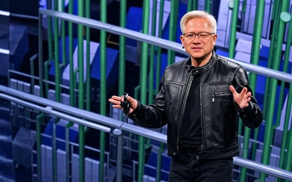 A person wearing a black leather jacket and dark clothing, standing on a stage with arms outstretched, presenting to an audience. The background features a modern, high-tech setting with green and blue structural elements, possibly representing a data center or technology exhibition, captured during a keynote or conference presentation.