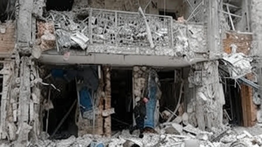 A heavily damaged building with a collapsed balcony and debris scattered across the ground. The structure shows extensive destruction, with broken concrete pillars, twisted metal railings, and exposed interiors. A person in dark clothing stands amidst the rubble, near the entrance of the building, highlighting the aftermath of a significant explosion or attack."
