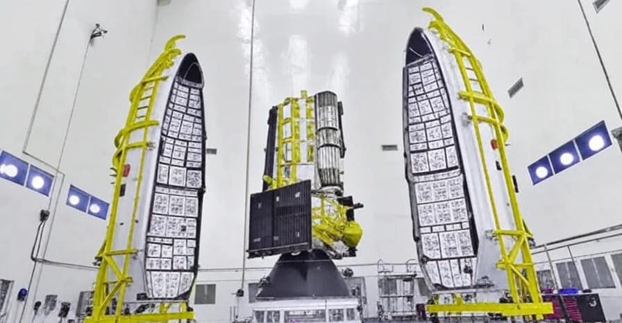 An image of the NASA-ISRO NISAR satellite assembly inside a cleanroom facility, featuring the satellite mounted on a launch vehicle with its protective fairings open, supported by yellow scaffolding, and surrounded by a high-tech workspace with lighting and equipment.
