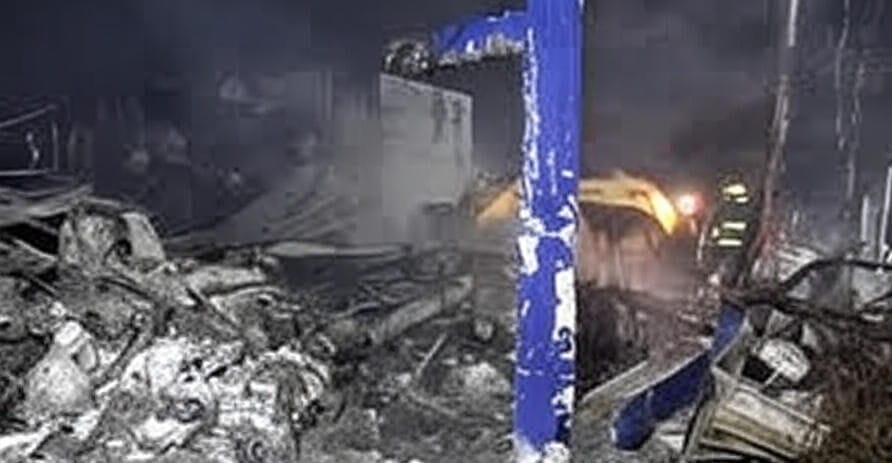 A nighttime scene of a devastated industrial site, with a pile of twisted metal, debris, and rubble illuminated by the headlights of a yellow excavator. Thick smoke rises from the wreckage, and a blue structural beam stands amidst the destruction. Emergency lights and personnel are visible in the background, indicating ongoing rescue or recovery efforts.