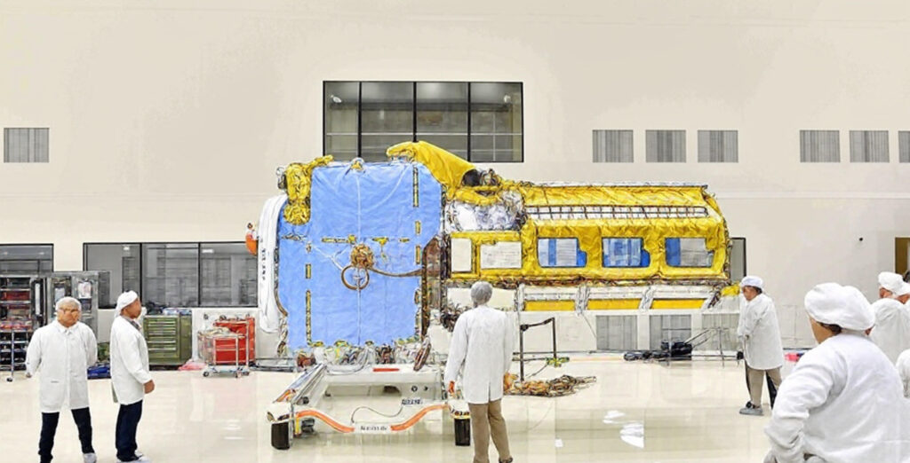 A group of technicians in cleanroom suits working on a large spacecraft or satellite with blue and yellow insulation panels inside a spacious, sterile facility.