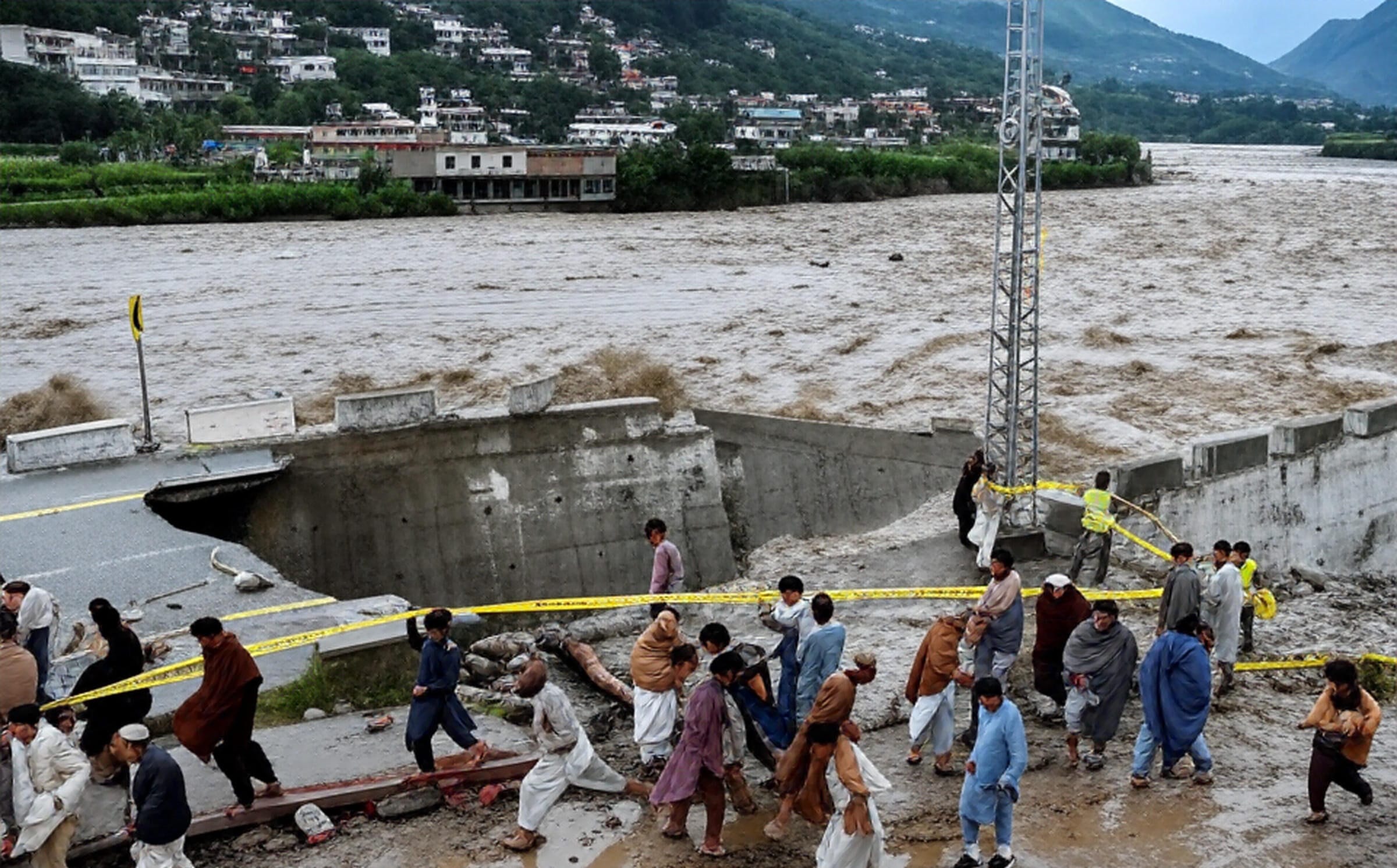 A group of people, including emergency personnel in high-visibility vests, work together near a damaged concrete barrier along a flooded riverbank. The swollen river, carrying muddy brown water, overflows its banks, threatening nearby buildings and a hillside town in the background. Yellow caution tape is strung across the area, and debris is scattered around as individuals assist each other in the muddy terrain, with mountains visible in the distance under a cloudy sky.