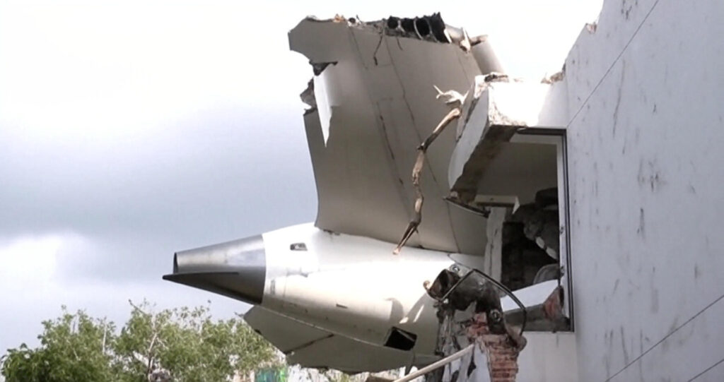 The wreckage of a jet aircraft embedded in the damaged upper section of a building, with a pointed nose and broken wing protruding from a shattered wall, surrounded by green trees under a clear blue sky, depicting the aftermath of the Bangladesh Air Force jet crash on July 21, 2025.