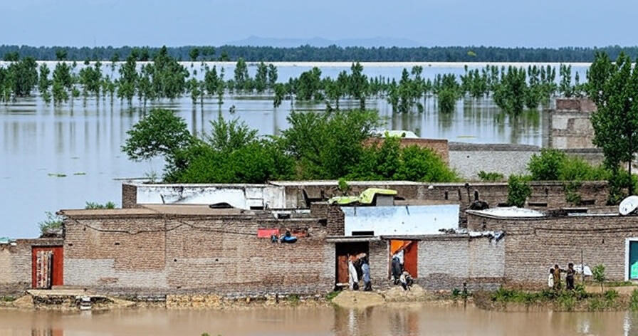 A row of brick houses with flat roofs stands partially surrounded by muddy floodwater, with a few people standing near the doors and windows. The water extends into the distance, submerging trees and vegetation, creating a vast reflective surface under a clear blue sky. In the background, a line of trees emerges from the flooded landscape, with hills faintly visible on the horizon, illustrating the extensive impact of flooding on the area.