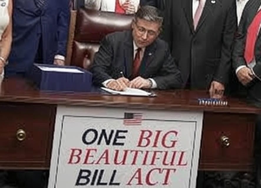A person in a suit signing a document at a desk, with a sign reading 'ONE BIG BEAUTIFUL BILL ACT' featuring an American flag, surrounded by other individuals in formal attire standing nearby.