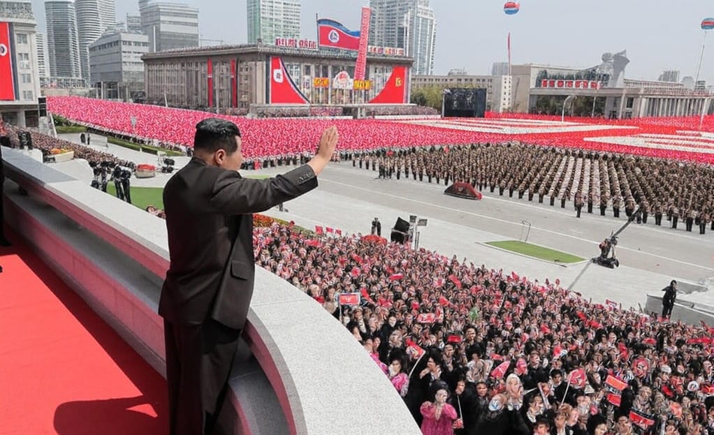 "Kim Jong-un waves to a massive crowd during a Pyongyang ceremony, with military troops and citizens waving flags, set against a backdrop of national banners and city skyline."