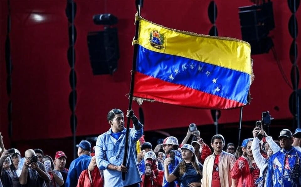 A man holding the Venezuelan flag before a cheering crowd, symbolizing national pride amid U.S. tensions.