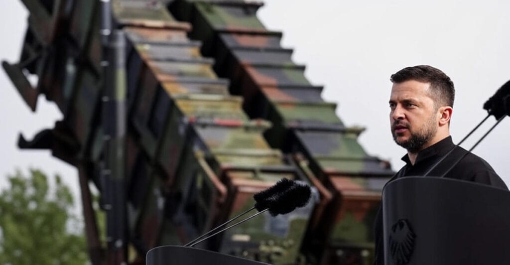 krainian President Volodymyr Zelenskyy, in a black suit, stands at a podium with microphones, speaking against a backdrop of a camouflaged military missile launcher, representing Ukraine's defense efforts amid the ongoing war."