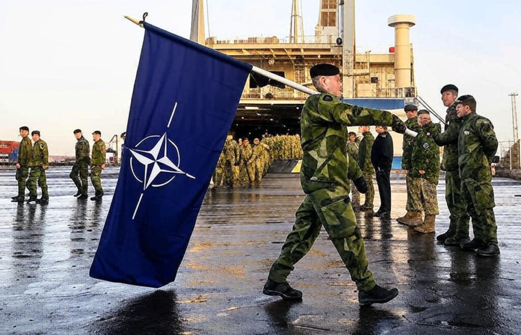 A soldier in camouflage uniform carries a large NATO flag with a white compass rose emblem on a blue background, walking on a wet surface in front of a military ship. Other soldiers in similar uniforms stand in formation, with additional personnel in the background near the ship's entrance, under a clear sky.