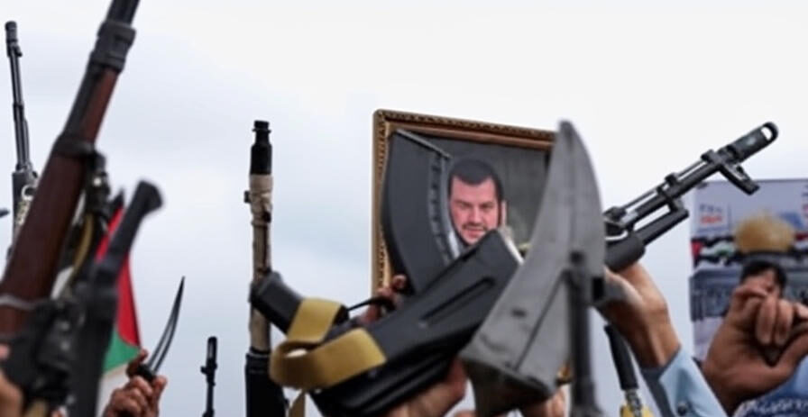 The image depicts a group of individuals holding various rifles and a large framed portrait of a man, raised aloft during what appears to be a public gathering or demonstration. The scene includes a mix of traditional and modern weaponry, with some rifles adorned with green and red flags. The background features a cloudy sky and a banner, suggesting a significant event or commemoration. The focus keyword "Israel" is not directly indicated in the image content.