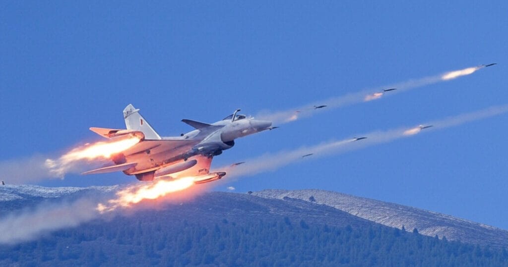 A military fighter jet in flight against a clear blue sky, launching multiple missiles with visible trails of smoke and fire. The jet is positioned in the foreground, angled upward, with the missiles streaking ahead. Snow-covered mountains are visible in the background, adding a dramatic effect to the scene.