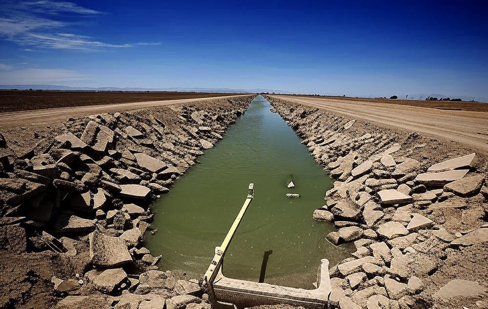 The image shows a narrow, man-made irrigation canal running through a dry, arid landscape under a clear blue sky. The canal is lined with rough, rocky embankments and contains greenish water, with a yellow measuring device or pipe extending across it. The surrounding area features dirt roads and barren fields, with distant mountains visible on the horizon, highlighting the scarcity of water in the region.