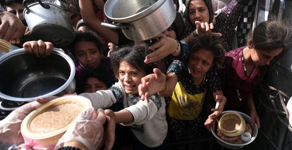 **Alt Text:** A group of children and adults reaching out with metal pots and bowls to receive food from a person wearing gloves, holding a container of food. The scene captures a crowded, desperate effort to obtain aid, highlighting a humanitarian crisis in a conflict-affected area.