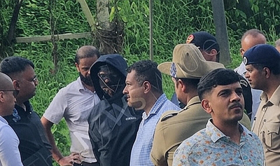 A group of police officers and civilians, including a suspect with a covered face, gathered at a lush green site during the Mangaluru Temple Case investigation in August 2025, highlighting the ongoing probe into alleged mass burials and cover-ups in Dharmasthala, Karnataka.