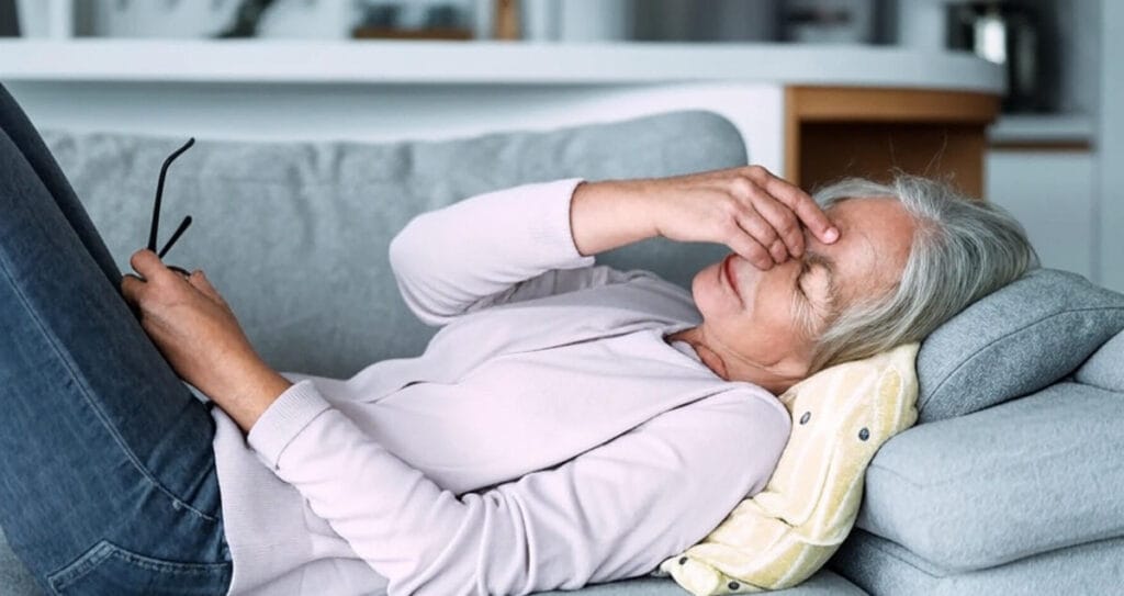 The image shows an elderly person lying on a gray couch, appearing to rest or experience discomfort. The individual is dressed in a light pink long-sleeve shirt and blue jeans, with one hand holding a pair of glasses and the other resting on their forehead. A yellow patterned pillow supports their head, and a light blue blanket is draped over their lower body. The background features a modern indoor setting with a kitchen counter and shelves, suggesting a home environment.