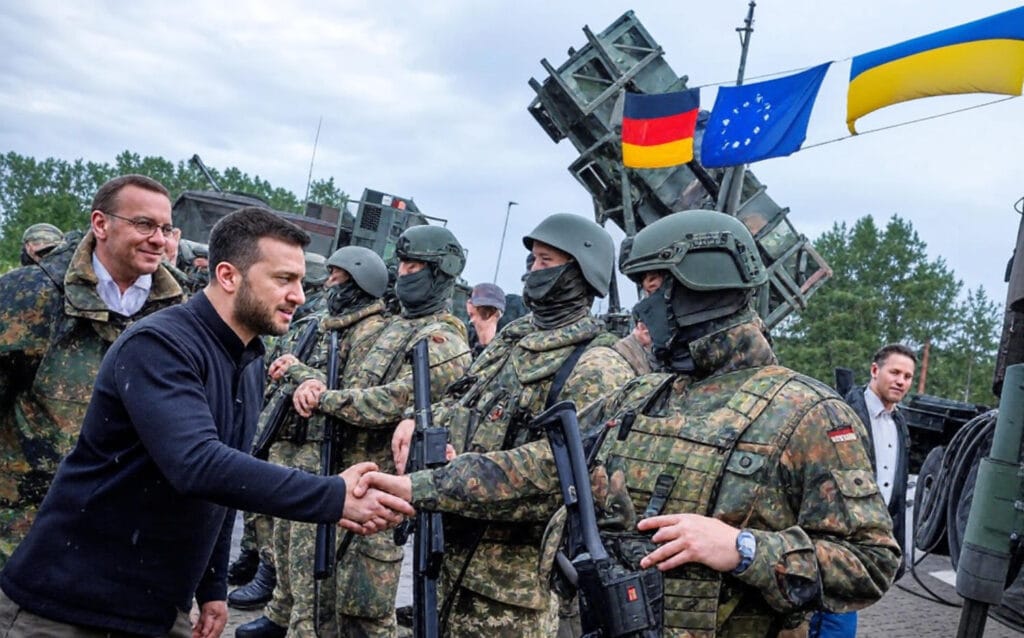 **Alt Text:** Ukrainian and German leaders shaking hands with soldiers, surrounded by military equipment and flags of Ukraine, Germany, and the EU, symbolizing international military support and cooperation.