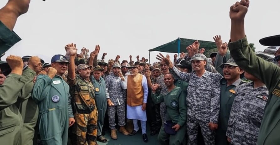 A group of military personnel in camouflage and green uniforms raise their fists in celebration, surrounding a central figure in an orange vest and white outfit, during the Operation Sindoor event, with a green canopy and clear sky in the background.