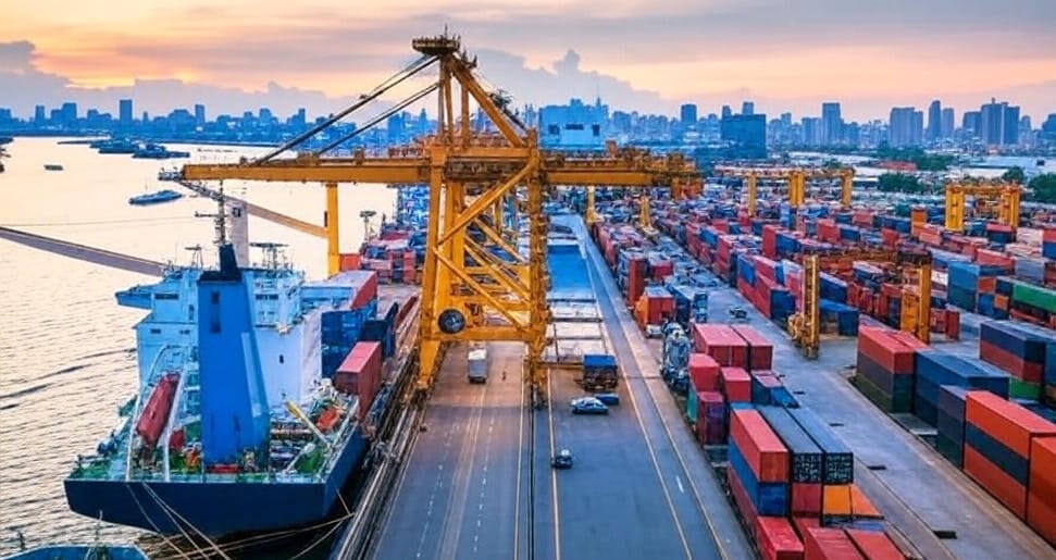 The image depicts an aerial view of a bustling port during sunset, featuring a large cargo ship docked alongside a pier. The ship is being unloaded by a prominent yellow crane, with numerous colorful shipping containers stacked in orderly rows along the dock. Additional cranes and trucks are visible, indicating active cargo handling. In the background, a city skyline with modern buildings stretches along the horizon, while the water reflects the warm hues of the setting sun, adding a vibrant glow to the scene.