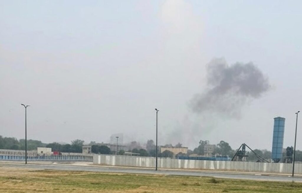 The image depicts a serene landscape featuring a calm lake surrounded by lush green trees under a bright blue sky with scattered white clouds. A wooden bench is positioned near the water's edge, accompanied by a tall, cylindrical structure. This scene contrasts with an earlier image showing smoke rising over a fenced industrial area with a road and streetlights, which may indicate a possible drone strike or related incident.