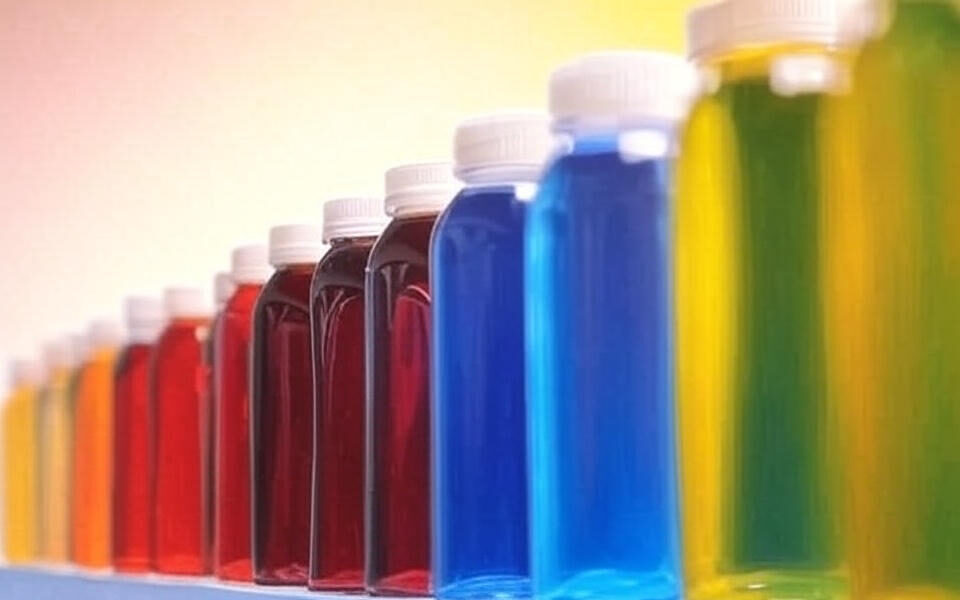 A row of plastic bottles filled with vibrant liquids in colors including red, blue, yellow, and green, representing artificial dyes used in food production, set against a blurred background.