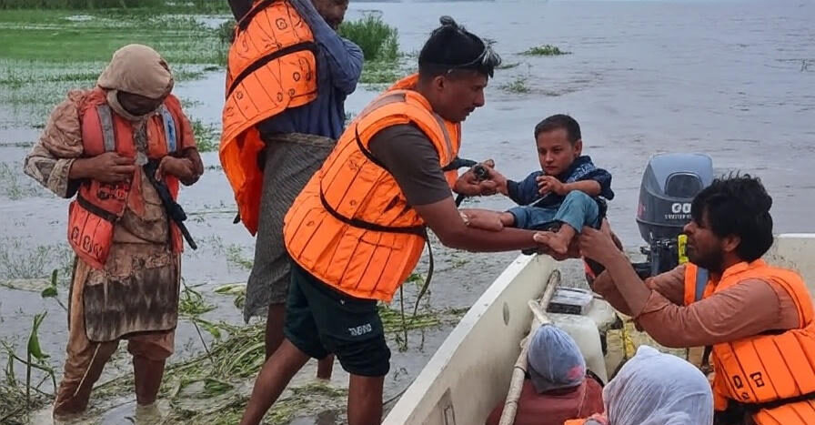 Rescue workers in orange life vests assisting a child and others from a flooded area, with a boat and water in the background.