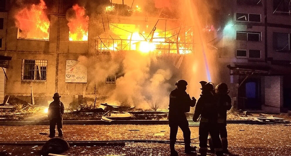 Firefighters working to extinguish a large fire engulfing a multi-story building at night in Ukraine, with flames and smoke rising from the structure, debris scattered on the ground, and water being sprayed to control the blaze.