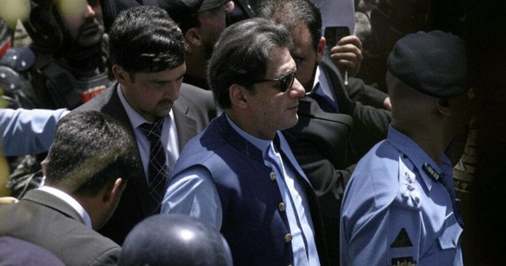 Imran Khan, wearing a blue traditional vest and sunglasses, is surrounded by security personnel and officials, including police officers and individuals in suits, as he moves through a crowded area with helmets and uniforms visible in the background.