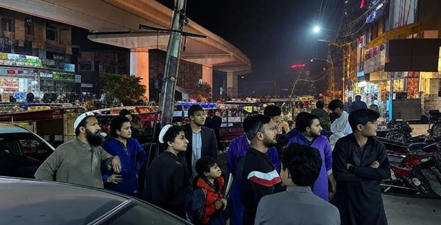 The image depicts a group of people standing on a busy urban street at night, looking upward with concern, likely in response to an earthquake. The scene includes a variety of individuals, including a child, surrounded by cars, rickshaws, and motorcycles, with a brightly lit overpass and commercial buildings in the background, illuminated by streetlights and shop signs.