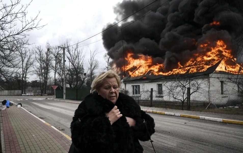 A woman in a black fur coat stands on a sidewalk with her hand to her chest, appearing distressed. Behind her, a house is engulfed in flames and thick black smoke, with fire consuming the roof. The scene takes place on a quiet street with a paved sidewalk, leafless trees, and a cloudy sky.