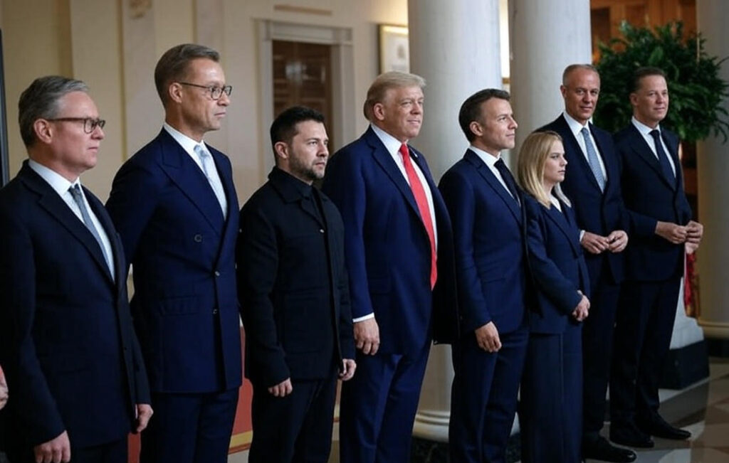 A line of world leaders, including U.S. President Donald Trump in a blue suit with a red tie, Ukrainian President Volodymyr Zelenskyy in a black outfit, and European leaders in formal suits, standing in the White House with columns and a polished floor, symbolizing a diplomatic gathering."