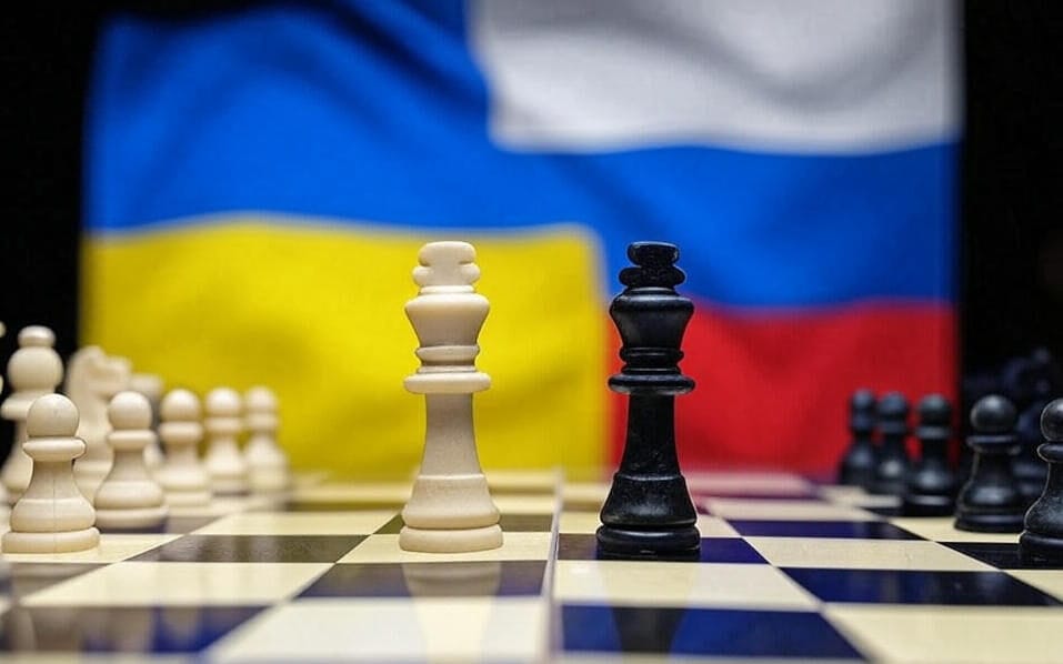 A chessboard with a white king and a black king positioned prominently in the center, facing each other. The background features the Ukrainian flag on the left with blue and yellow colors, and the Russian flag on the right with red, white, and blue colors. The chess pieces are arranged in their starting positions, with white pieces on the left and black pieces on the right.