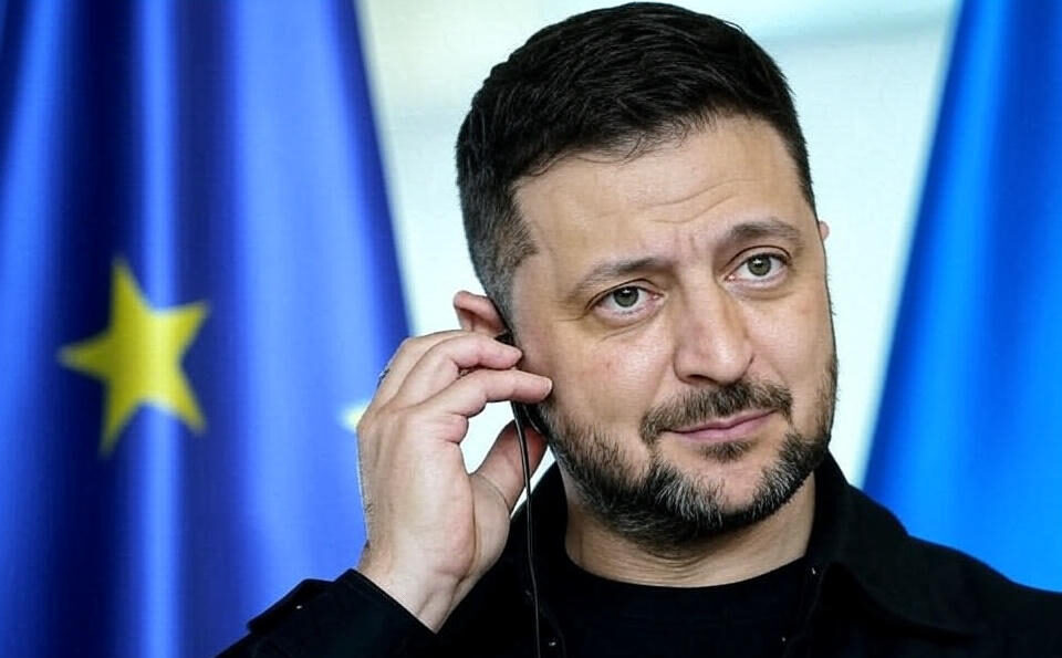 Ukrainian President Volodymyr Zelensky adjusting an earpiece, standing in front of European Union flags with yellow stars on a blue background, dressed in a black outfit.