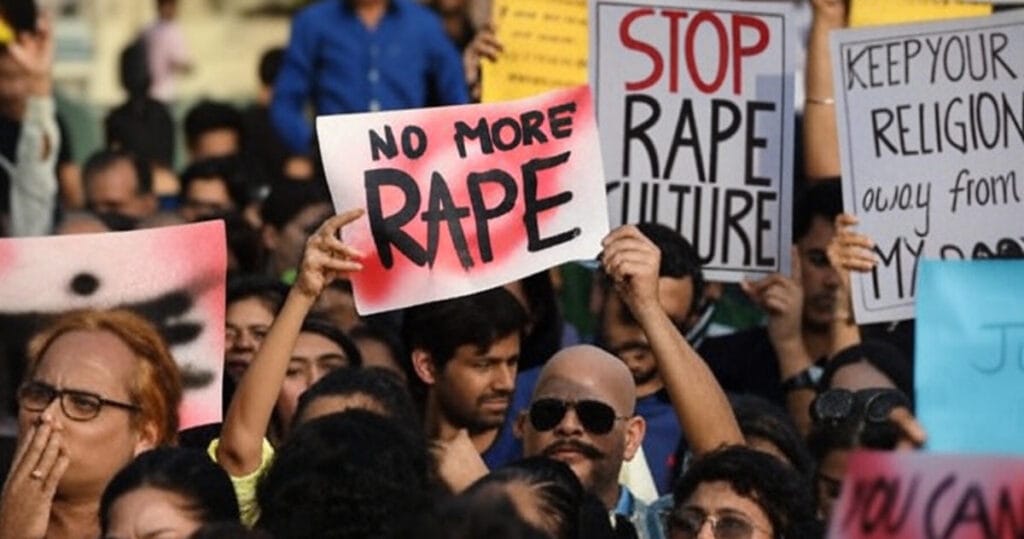 A large crowd protests against sexual violence in India, holding signs reading 'NO MORE RAPE,' 'STOP RAPE CULTURE,' and 'KEEP YOUR RELIGION AWAY FROM MY BODY,' reflecting public outrage following cases like the Mangaluru Temple Case and the Kolkata doctor rape and murder incident in 2024-2025.
