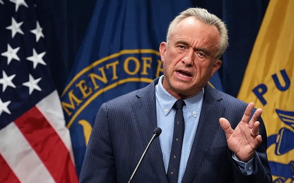 "A U.S. official in a suit speaking at a podium with a microphone, standing in front of the U.S. flag and a banner with the 'Department of' seal, gesturing with a raised hand during a formal address."