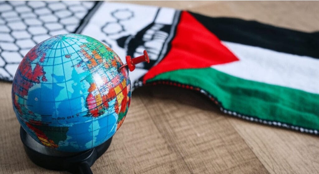 A small globe with a red pin marking a location, placed on a wooden surface with a magnifying glass beneath it. In the background, a fabric featuring the Palestinian flag with red, green, white, and black colors, along with a patterned design, symbolizes global attention on the region.
