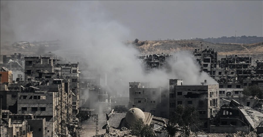 The image depicts a war-torn urban landscape with numerous damaged and partially collapsed buildings. Thick smoke rises from multiple locations, obscuring parts of the scene. Debris is scattered across the ground, and a prominent dome-shaped structure stands amidst the destruction. The background features barren hills under a hazy sky, emphasizing the desolate and chaotic atmosphere.