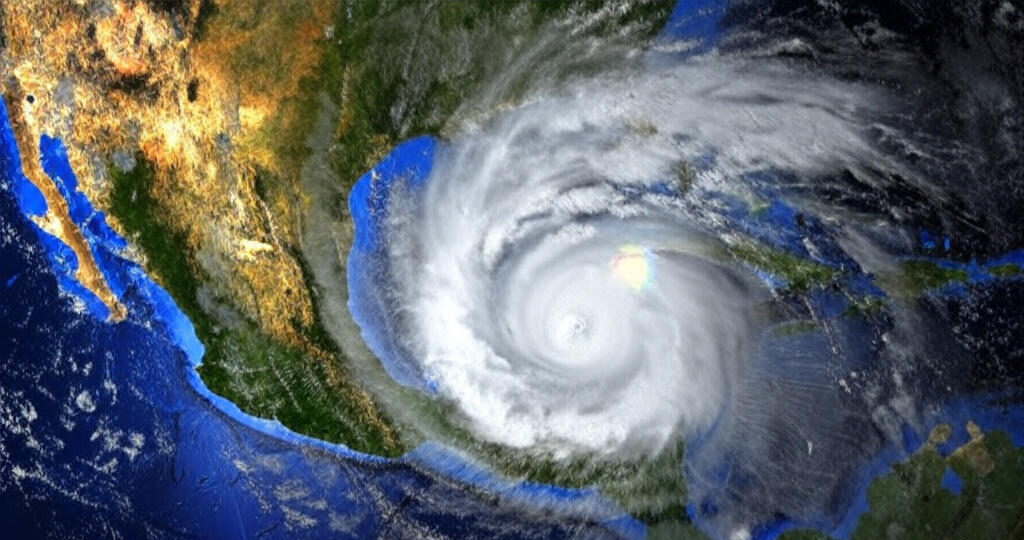 The image depicts a satellite view of a hurricane approaching the Gulf of Mexico, with its swirling cloud pattern prominently visible. The storm is positioned near the coastlines of Central America, Mexico, and parts of the southern United States, with the eye of the hurricane clearly defined. The surrounding landmasses are illuminated in shades of gold and green, contrasting with the dark blue oceans and the white and gray clouds of the storm.
