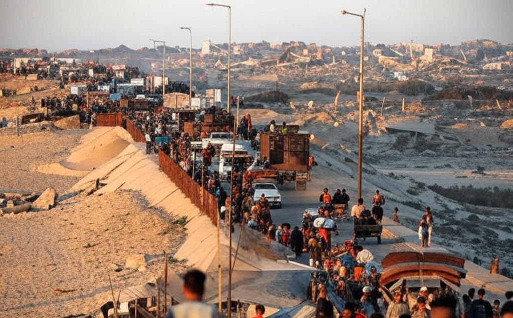 The image portrays a bustling border crossing scene in a desert landscape during golden hour lighting. A long line of pedestrians, vehicles, and carts extends along a fortified barrier wall topped with fencing, amid sandy dunes and scattered debris. In the background, a densely packed urban skyline of multi-story apartment buildings emerges through a hazy atmosphere, with some structures appearing damaged or under construction. Streetlights line the elevated roadway, and diverse groups of people—carrying belongings, pushing carts laden with goods, and traveling in trucks and taxis—navigate the passage, evoking a sense of migration and commerce in a rugged, arid environment.