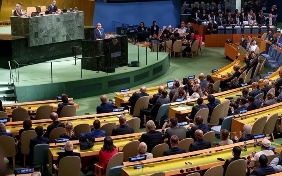 The image depicts an overhead view of the United Nations General Assembly hall during a session. A speaker stands at the central podium on a raised green platform, addressing the assembly. Rows of delegates are seated at curved desks with yellow tabletops, equipped with microphones and nameplates indicating countries such as "Canada." The audience includes individuals in formal attire, some applauding or engaged in discussions, with a presiding panel visible on an elevated marble dais to the left. The hall features a distinctive green carpet and blue accents in the background.